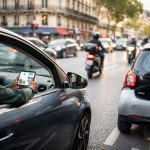 Rue d’une grande ville en 2026, voitures en circulation dense et citadine garée, un conducteur compare ses dépenses automobiles sur son smartphone.