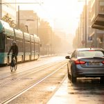 Scène de mobilité urbaine : un cycliste circule près d’un tramway dans une grande ville, avec une voiture en autopartage au bord de la route.