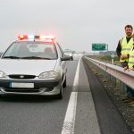 Une voiture immobilisée sur une bande d’arrêt d’urgence, feux de détresse allumés, avec un homme et une femme en gilets jaunes derrière une glissière de sécurité.